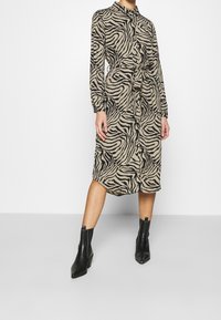 Šaty se zebrovaným vzorem v béžové a černé barvě, s límečkem, dlouhými rukávy, páskem v oblasti pasu a délka po kotníky, doplněné černými kotníkovými botami.