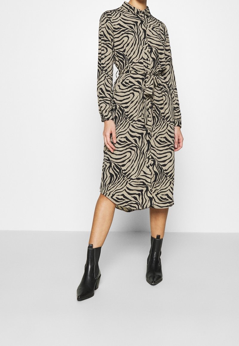 Šaty se zebrovaným vzorem v béžové a černé barvě, s límečkem, dlouhými rukávy, páskem v oblasti pasu a délka po kotníky, doplněné černými kotníkovými botami.