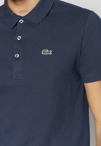 Polo en coton bleu marine avec un col à trois boutons, un tissu texturé et un petit logo de crocodile brodé en vert sur la poitrine.