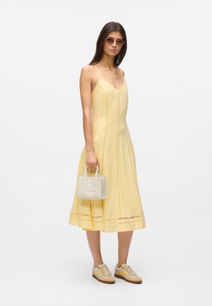 Kobieta w żółtej, bezrękawnikowej sukience midi, w butach sportowych i okularach przeciwsłonecznych, trzymająca małą białą torebkę na tle jednolitego tła.