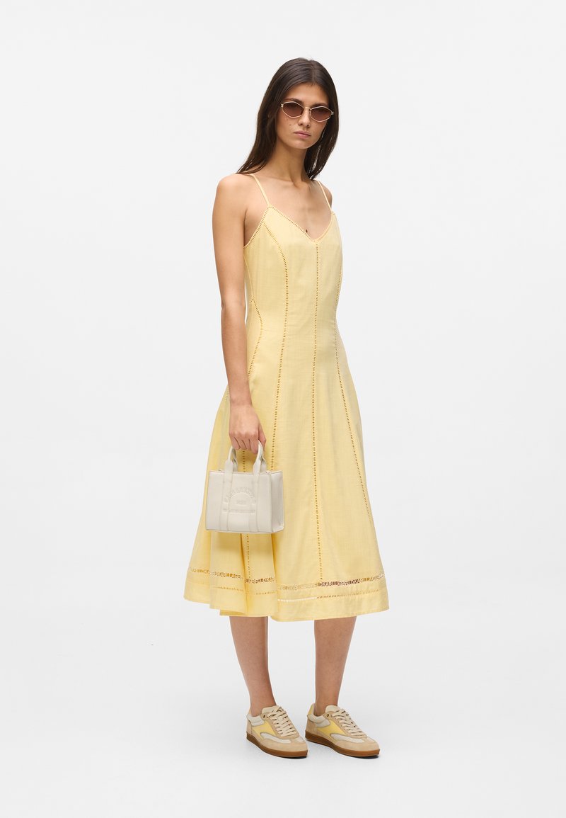 Γυναίκα που φοράει ένα κίτρινο μιντι φόρεμα χωρίς μανίκια, αθλητικά παπούτσια και γυαλιά ηλίου, κρατώντας μια μικρή λευκή τσάντα σε απλό φόντο.