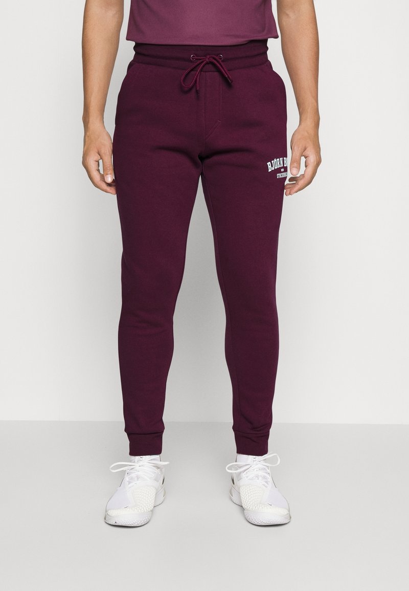 Man i burgundoröda joggerbyxor med vitt märke på låret och vita sportskor, stående mot en enkel ljus bakgrund.