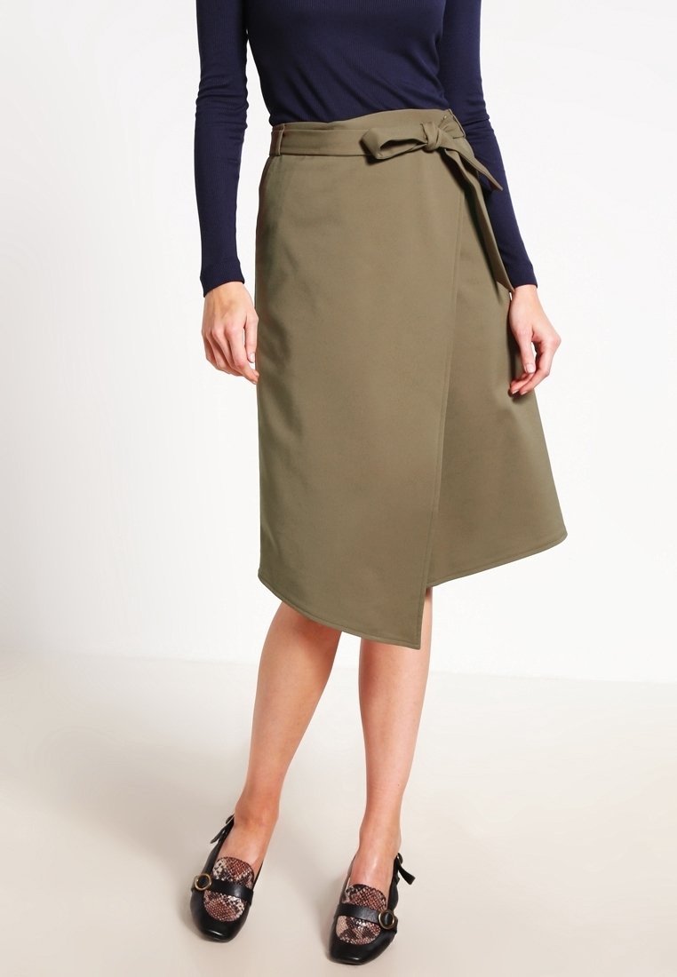 Femme portant une jupe portefeuille olive longueur genou avec une ceinture nouée et des chaussures noires avec un détail imprimé serpent.
