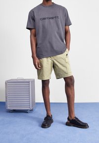 Grijze oversized t-shirt met "carhartt." opdruk, khaki cargoshorts met zijzakken en zwarte leren sandalen. Zilveren rolkoffer op de achtergrond.