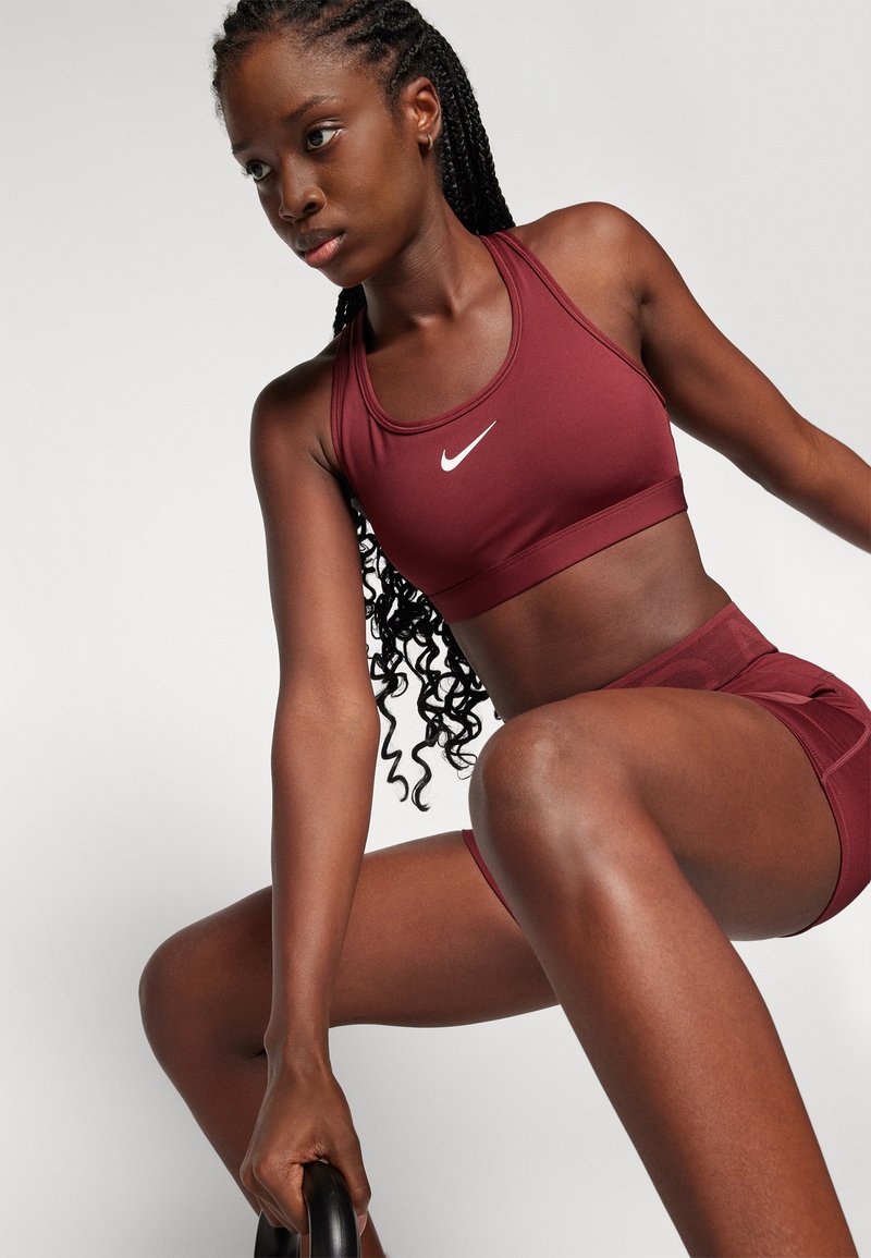 Reggiseno sportivo borgogna con logo Nike bianco, abbinato a pantaloni corti borgogna. Presenta una texture liscia e un design aderente, adatto per l'abbigliamento sportivo.