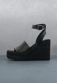 Sandale compensée noire avec semelle plateforme tressée, large bride perforée à l'avant, et bride de cheville réglable avec boucle argentée sur fond gris.