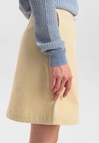Jupe en velours côtelé beige de forme droite, avec des nervures subtiles, des poches sur les côtés et un ourlet atteignant le milieu de la cuisse. Portée avec un pull texturé bleu.