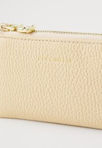 Beige lærpung med teksturert overflate, gullglidder og preget merke «Coccinelle» i gull. Kompakt design med en slank silhuett.