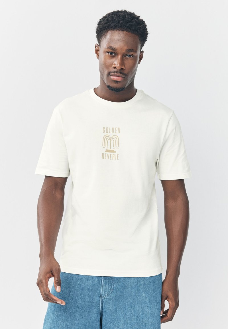 Jonge man draagt een wit T-shirt met de tekst "GOLDEN REVERIE" en afbeelding, gecombineerd met blauwe spijkershorts, staand tegen een effen achtergrond.