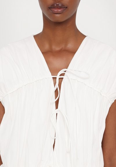 White, lightweight fabric garment with a deep V-neck and gathered detailing. Features a tie closure at the neckline and short sleeves.