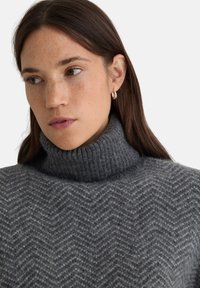 Maglione grigio a collo alto lavorato a maglia con motivo a spina di pesce, dotato di collo a coste e una morbida consistenza, abbinato a piccoli orecchini a cerchio.