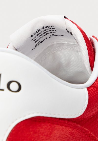 Κόκκινο και άσπρο αθλητικό παπούτσι με υφασμάτινη κόκκινη επένδυση και λεία λευκή δερμάτινη λεπτομέρεια. Η εσωτερική ετικέτα περιέχει εκτυπωμένο κείμενο σε μαύρο χρώμα.