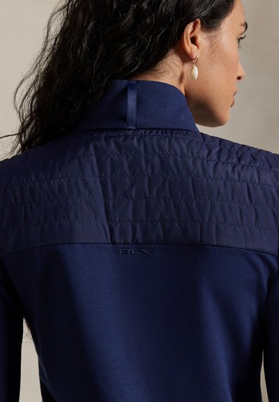 Σκούρο μπλε μπουφάν με καπιτονέ μπροστινό πάνω μέρος, ομαλή υφή στο κάτω μέρος και ανάγλυφο λογότυπο RLX. Δομημένος γιακάς με θηλιά κρεμάσματος.