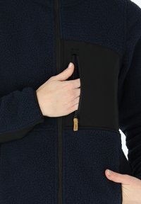 Navy fleece jacket with a black panel, featuring a zippered chest pocket and a soft texture. Includes black cuffs and a full zipper.