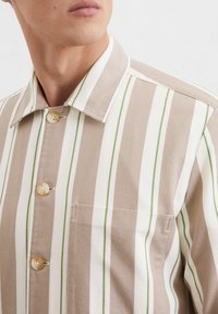 Gestreept shirt in beige en wit met groene accenten; heeft een puntige kraag, knoopsluiting en een borstzak. Gestructureerde stof.