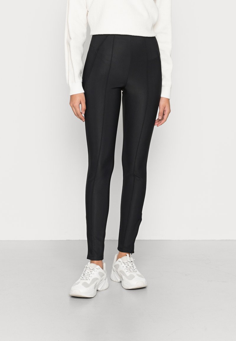 Femme portant un pantalon noir ajusté, un pull blanc et des baskets blanches épaisses debout sur un sol gris clair contre un mur blanc.