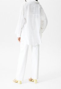 White linen shirt with dropped shoulders, and white wide-leg pants, paired with yellow block-heeled sandals. Textured fabric and relaxed design.