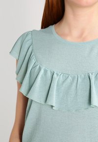 Ljusgrön topp med rund halsringning, som har en texturerad tyg och lager av volangdetaljer på ärmarna och bröstområdet.