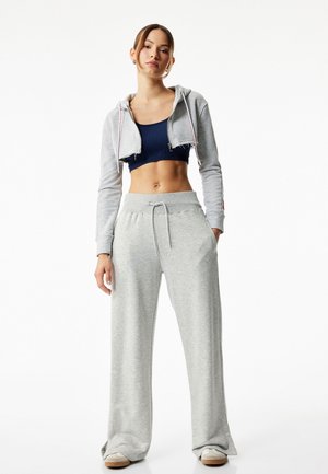 Grijze cropped hoodie met witte en rode accenten, gecombineerd met lichtgrijze wijde sweatpants. Donkerblauwe crop top eronder. Glad, casual stof.