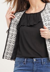 Top negro con volantes debajo de una chaqueta texturizada en blanco y negro. Pantalones vaqueros de denim visibles, que muestran un diseño ajustado con un detalle de botón.