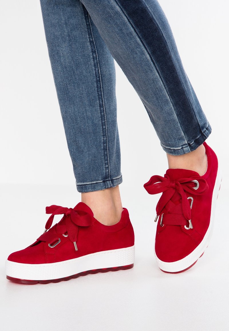 Baskets en daim rouge avec une semelle en caoutchouc blanche, dotées de larges lacets rouges et œillets. Un jean à dégradé bleu en denim complète le look.