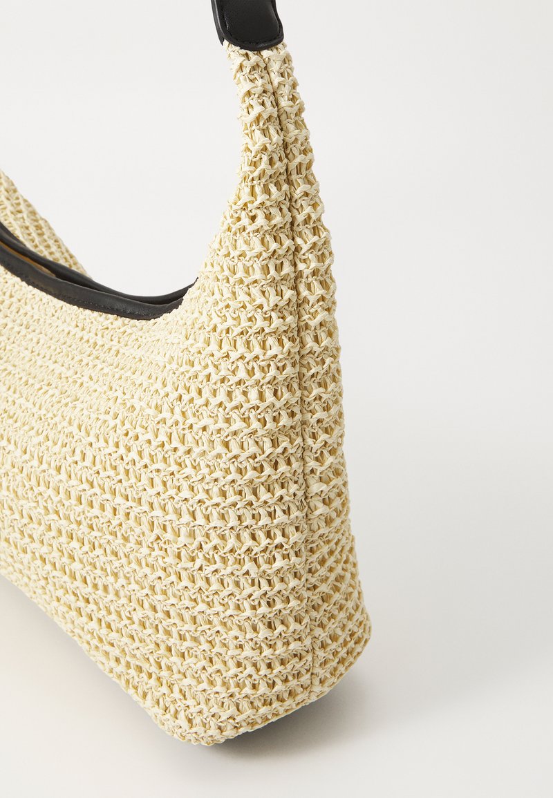 Bolso de paja tejido de color crema, con un asa de cuero negro y una forma redondeada, que muestra una superficie texturizada y costuras detalladas.