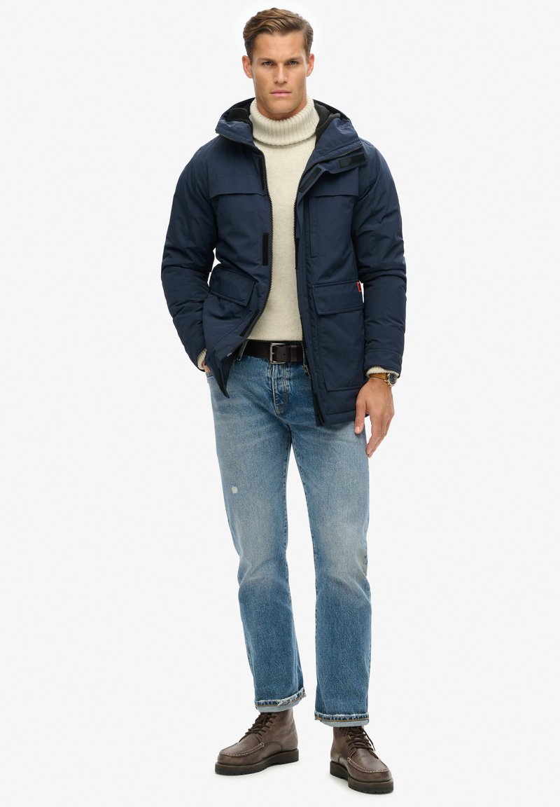 Marineblauwe pufferjack met capuchon, vier voorzakken en een ritssluiting, gedragen over een crèmekleurige turtleneck, gecombineerd met lichte spijkerbroek en bruine laarzen.