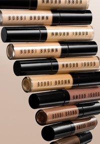 Mehrere Tuben Bobbi Brown Concealer in verschiedenen Beige- bis Brauntönen, diagonal auf einem hellbeigen Hintergrund angeordnet.