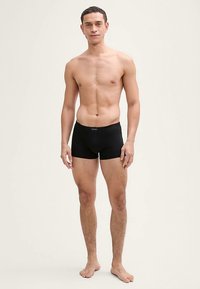 Svarta boxer shorts i tyg med slät textur, med en åtsittande design och ett elastiskt midjeband. Modellen visas utan skjorta.