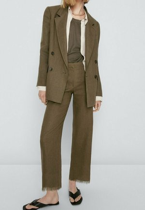 Femme porte un blazer marron croisé et un pantalon assorti avec des ourlets bruts, superposés à des hauts gris et blancs, associés à des sandales slides noires.