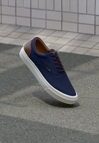 Marineblaue Sneaker mit weißer Sohle und braunem Lederfersenbereich, schwebend über einem strukturierten Pflaster und einem gefliesten Wandhintergrund.