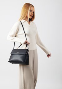 Black leather handbag with a structured shape, folded flap, and golden hardware accents. Model wearing a cream-colored sweater and beige pants.