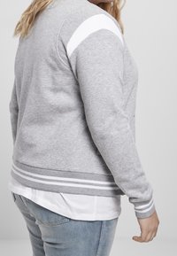 Sweat-shirt gris avec des rayures blanches sur les manches, poignets et ourlet côtelés. Tissu texturé, coupe ajustée, superposé à une chemise blanche.