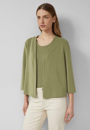 Frau trägt eine olivgrüne Strickjacke mit offenem Vorderteil und dreiviertel Ärmeln über einem passenden Oberteil und cremefarbenen Hosen.