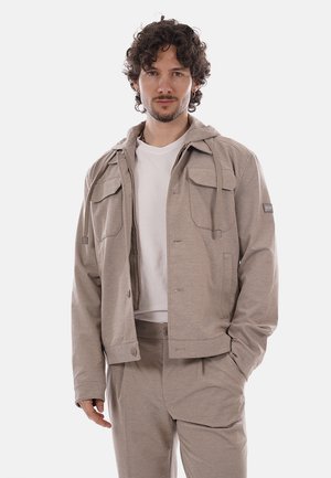 Uomo con capelli ricci che indossa una giacca beige sopra una camicia bianca e pantaloni beige coordinati, in piedi con una mano in tasca.