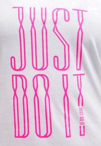 T-shirt bianco in cotone con testo rosa audace che dice "JUST DO IT" in un carattere moderno ed allungato, con una piccola stampa aggiuntiva.