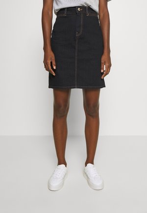 Person iført en sort jeansnederdel til knæet med kontrastsyninger, en stribet skjorte stukket ind, og hvide sneakers, stående på et gråt gulv.