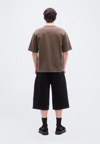 Άτομο που στέκεται γυρισμένο προς τα πίσω, φοράει ένα καφέ φαρδύ μπλουζάκι, μαύρα σορτς φαρδιάς γραμμής, μαύρες κάλτσες και μαύρα σανδάλια με αγκράφες, σε λευκό φόντο.