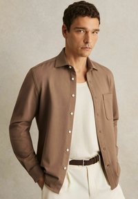 Cămașă lungă maro cu (mâneci) lungi, cu nasturi pe față, textură subtilă și un singur buzunar pe piept purtată peste un top crem cu dungi.