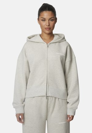 Donna che indossa una felpa con cappuccio grigio chiaro con zip e pantaloni della tuta abbinati con piccolo testo bianco su petto e coscia, in piedi su sfondo bianco.