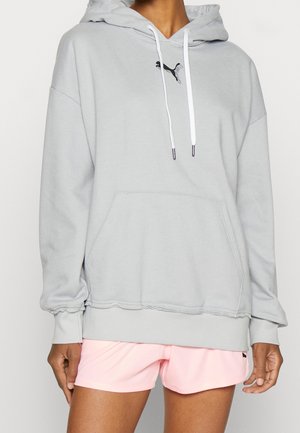 Personne portant un sweat à capuche Puma gris clair avec une poche avant et des cordons blancs, assorti à un short de sport rose clair.