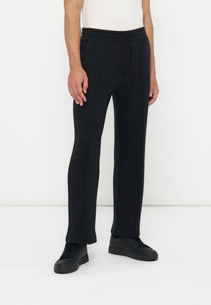 Mann trägt schwarze gerade geschnittene Hose mit Kordelzug und Seitentaschen sowie schwarze Sneaker, steht vor einer weißen Wand.