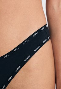 Crni tanga gaćice od mekog materijala s uskim pojasom, s ponavljajućim bijelim "GUESS" natpisima duž gornje ivice.