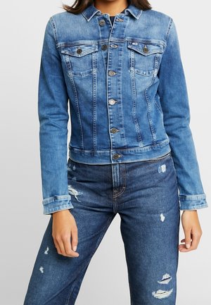 Femme portant une veste en jean bleu moyen avec des boutons en métal et un jean bleu foncé délavé, sur un fond gris uni.