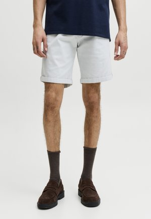 Mand iført lysegrå opsmøgede shorts, mørkebrune sokker, mørkebrune loafers og en mørk marineblå skjorte står mod en ensfarvet baggrund.