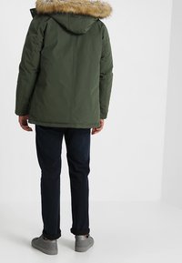 Veste isolante verte avec capuche doublée de fourrure, présentant une coupe droite, portée avec un jean foncé et des chaussures grises.