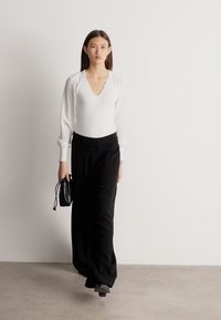 Γυναίκα που περπατά φορώντας λευκό μακρυμάνικο μπούστο με V λαιμόκοψη, μαύρο παντελόνι φαρδύ, μαύρα παπούτσια, κρατώντας μικρή μαύρη τσάντα μπροστά σε απλό ανοιχτόχρωμο τοίχο.