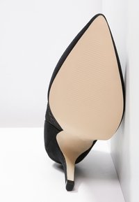 Scarpa nera in suede con tacco alto a punta e sottile tacco a stiletto. La suola è beige con texture rigata per una migliore presa.