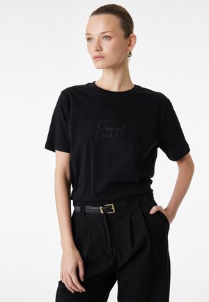 Femme portant un t-shirt noir "Jungle bird", un pantalon noir avec une ceinture noire et des boucles d'oreilles dorées, debout avec une main dans la poche.