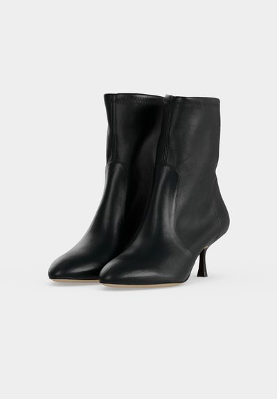 Paire de bottes mi-mollet en cuir noir avec bouts pointus et talons hauts fins, présentées sur un fond gris clair uni.